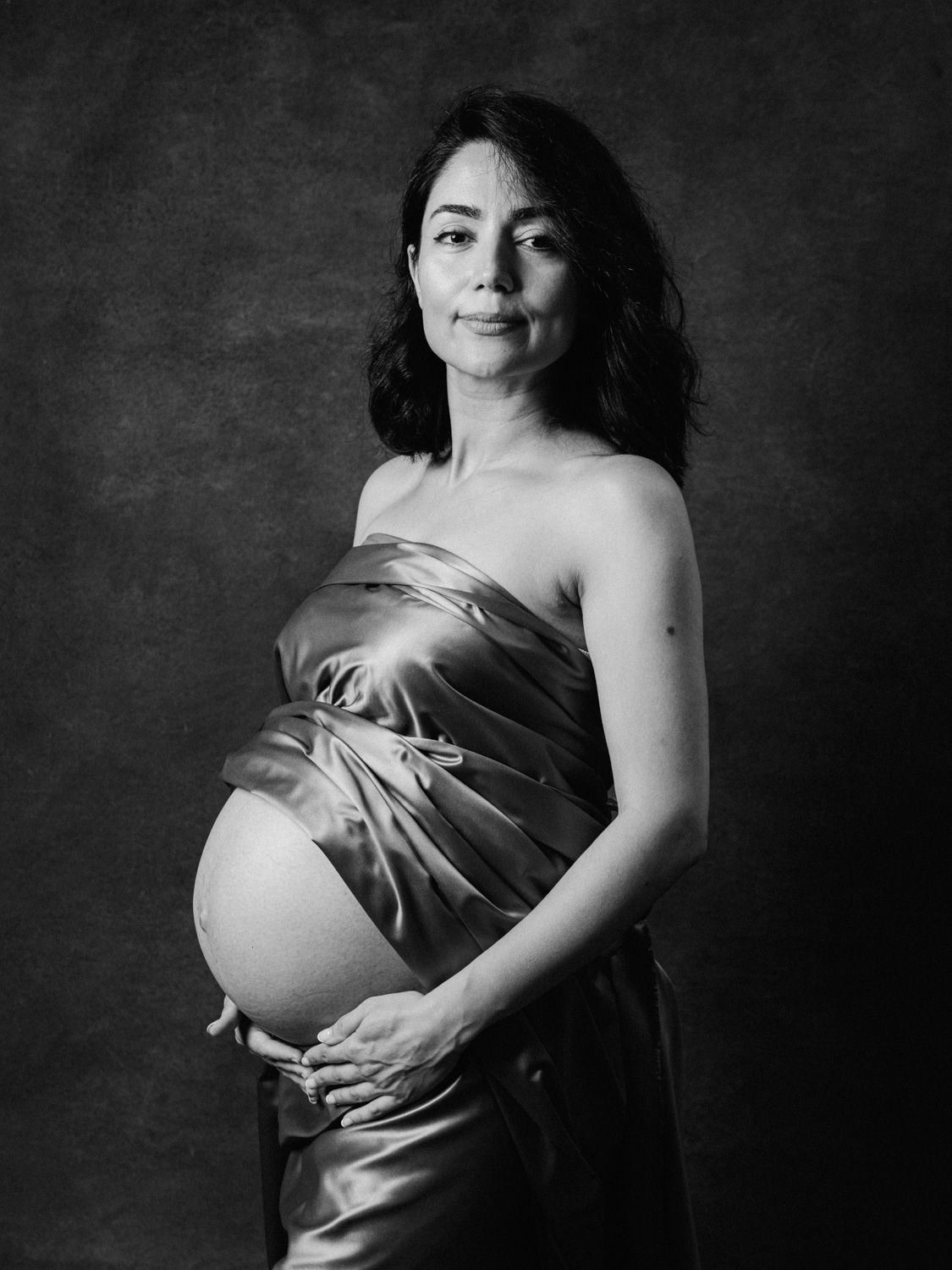 Black and white maternity portrait of pregnant woman in silk wrap gown cradling baby bump in studio.