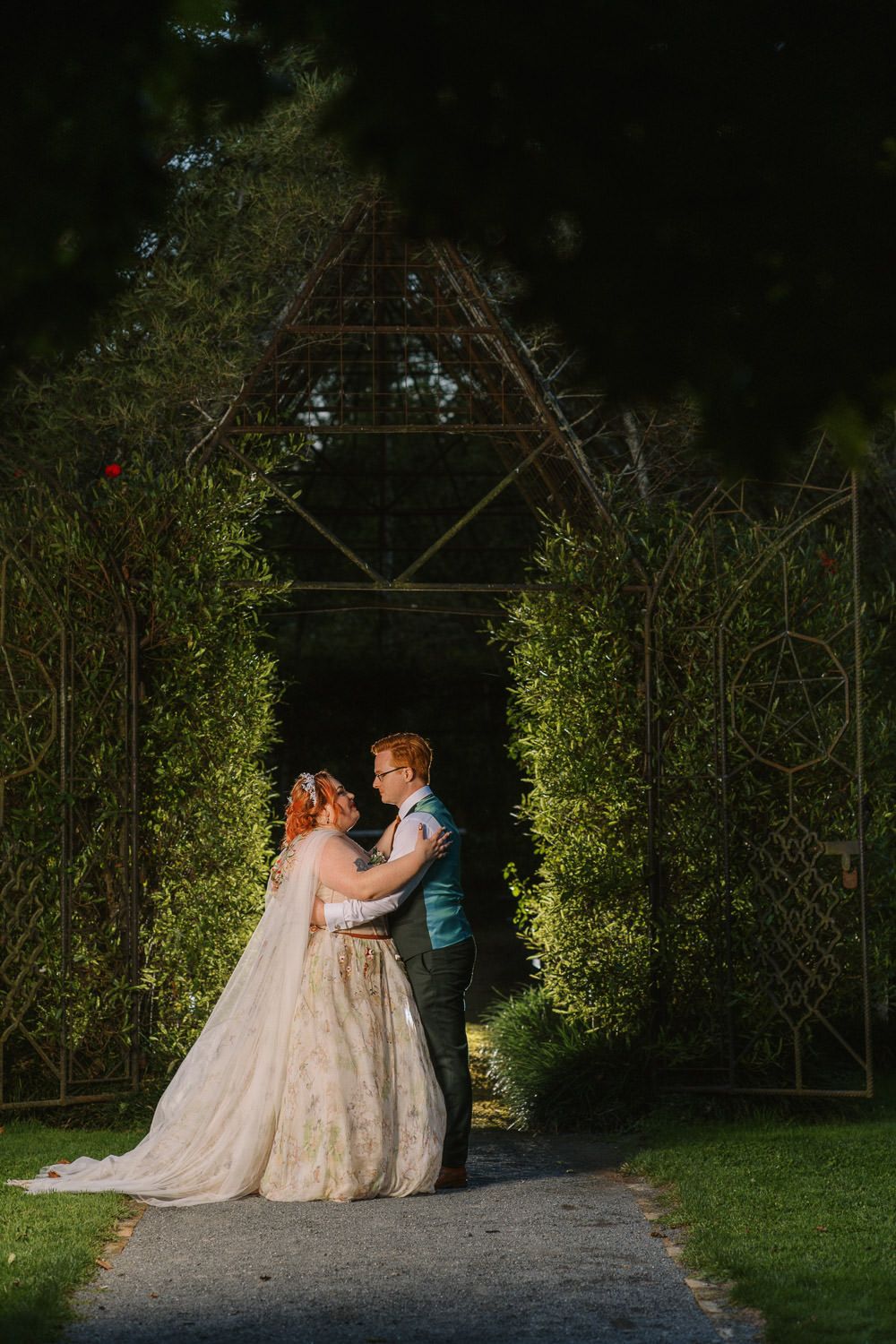 Bride and groom share a romantic moment under a garden arch surrounded by lush green hedges at golden hour.