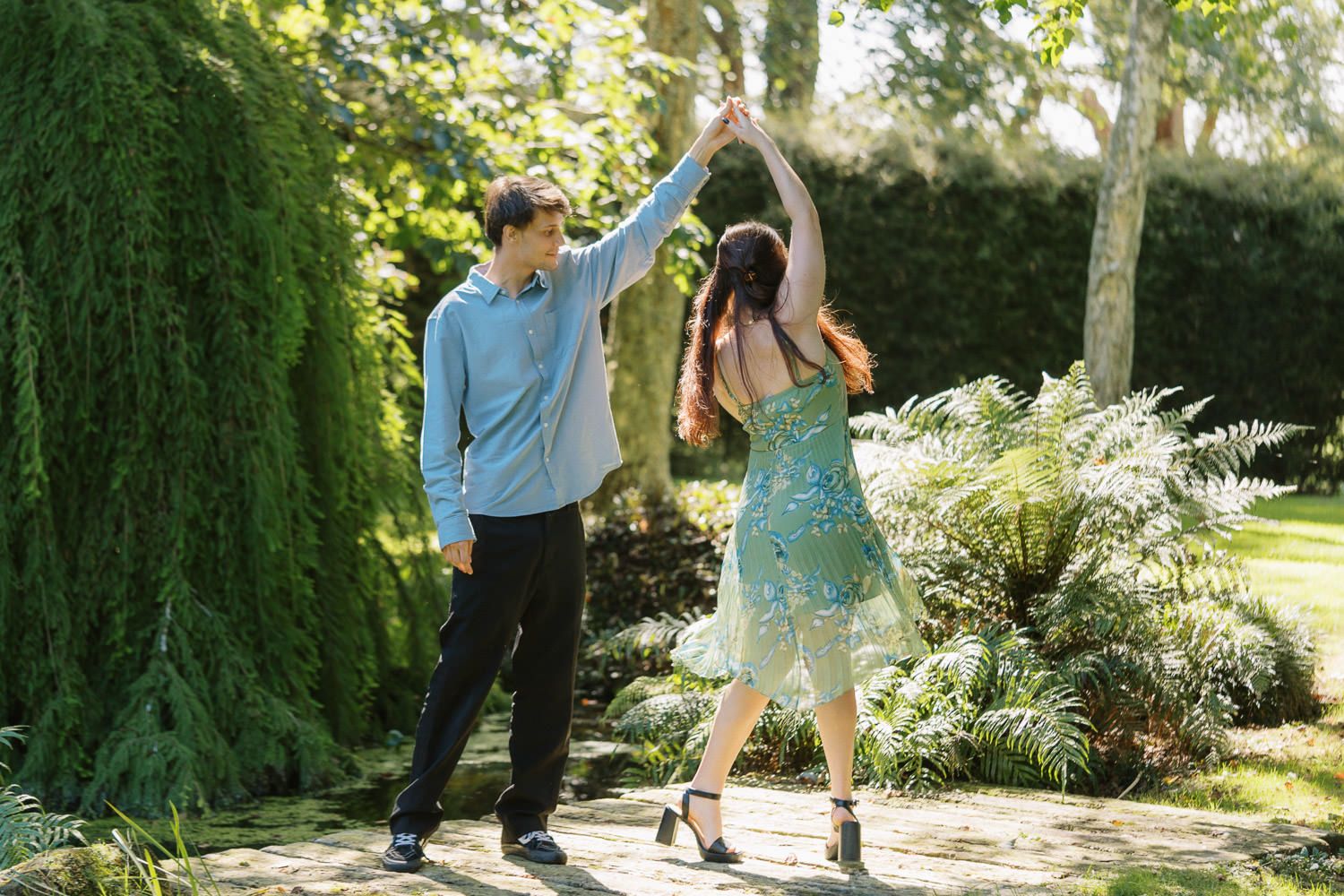 Couple dancing outdoors in a lush garden, man spinning woman in a blue dress on a stone path.