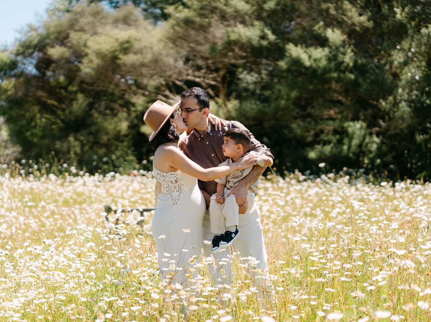 A couple sharing a kiss while holding their child in a sunny wildflower meadow surrounded by lush green trees.