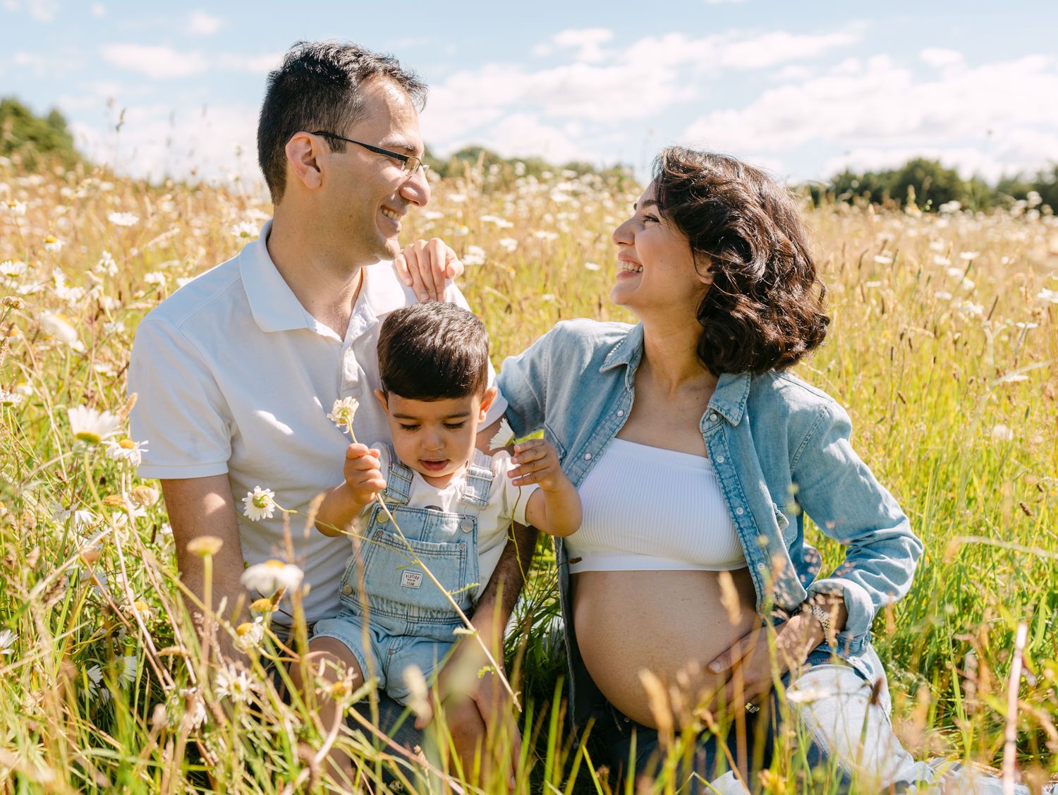 Pregnant woman and husband sit in flower field with toddler son on sunny day for maternity photo.