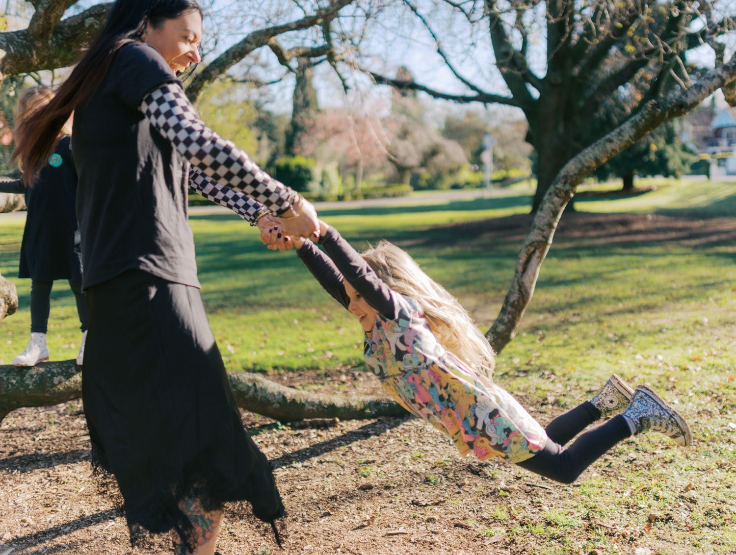 A woman swinging a young blonde girl by her hands in a green park on a sunny day.