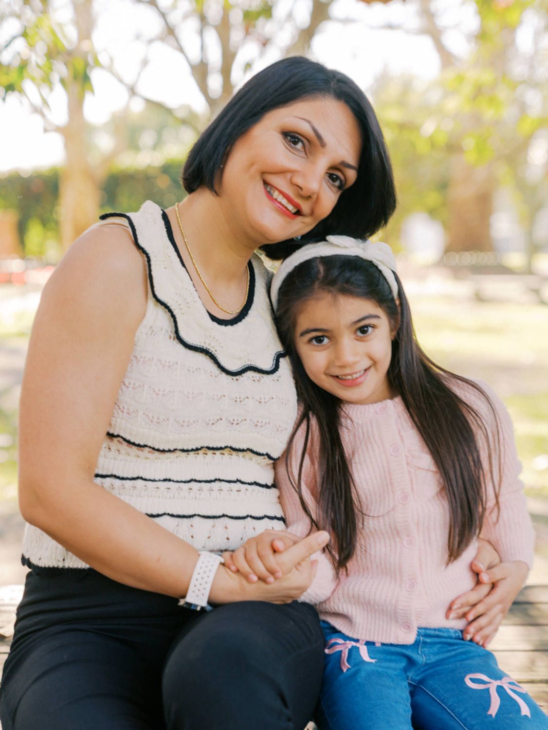 A smiling woman and young girl with dark hair sit together on a bench in a sunny park, wearing pink and white outfits.