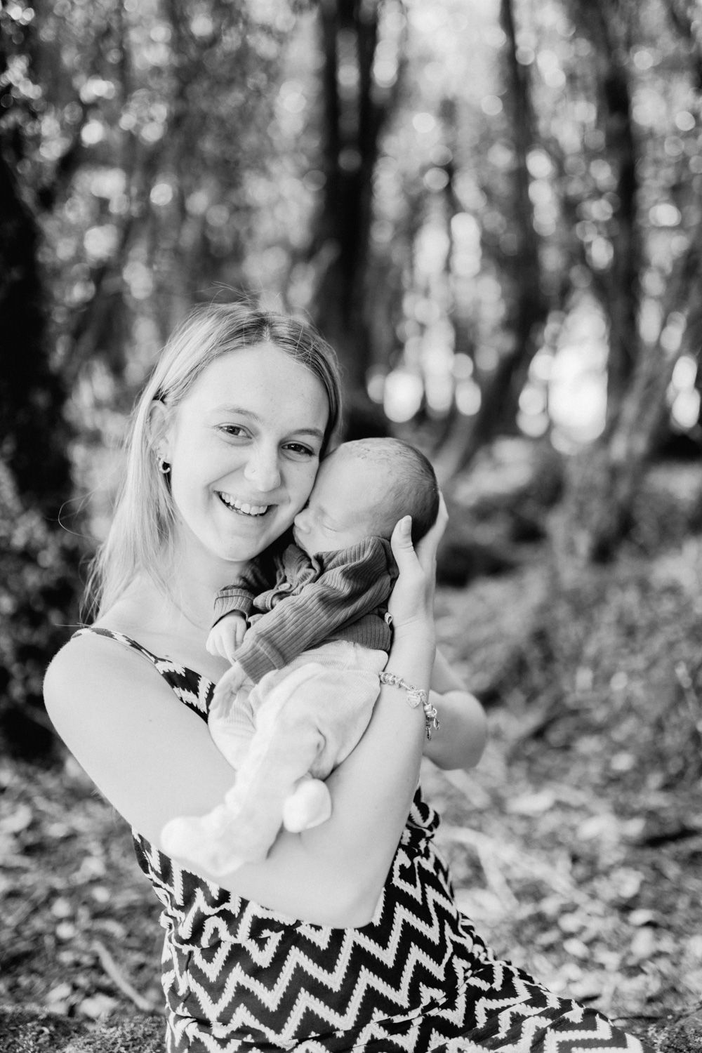 Black and white photo of a smiling blonde woman holding a newborn baby in a sunlit forest setting.