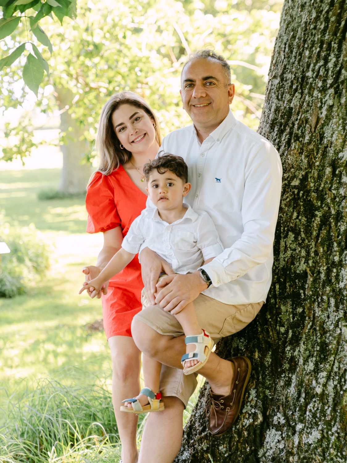 A smiling family of three poses by a tree, mom in red dress, dad holding their young son outdoors.