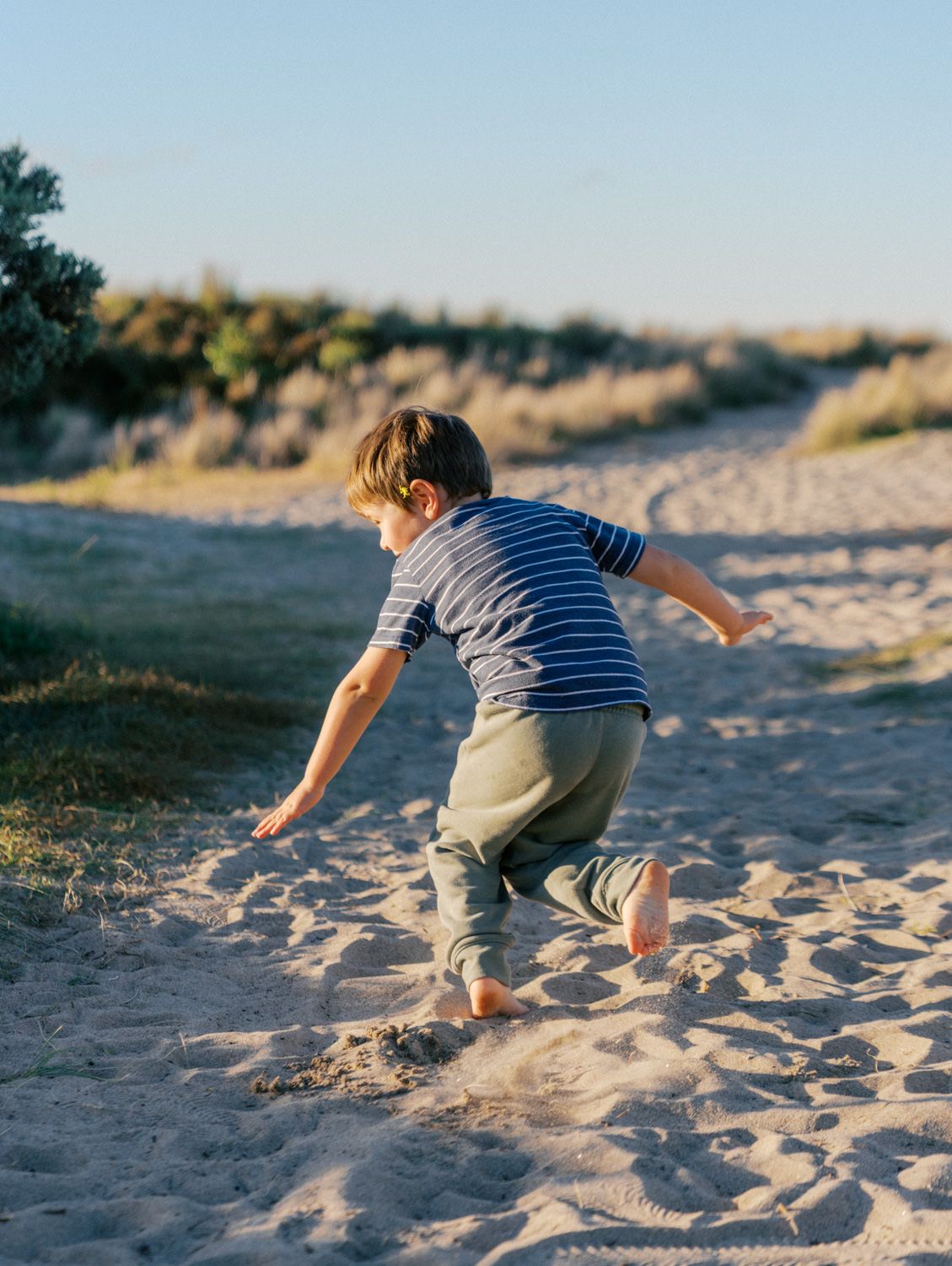A young boy in a striped shirt runs barefoot on sandy beach dunes during golden hour.