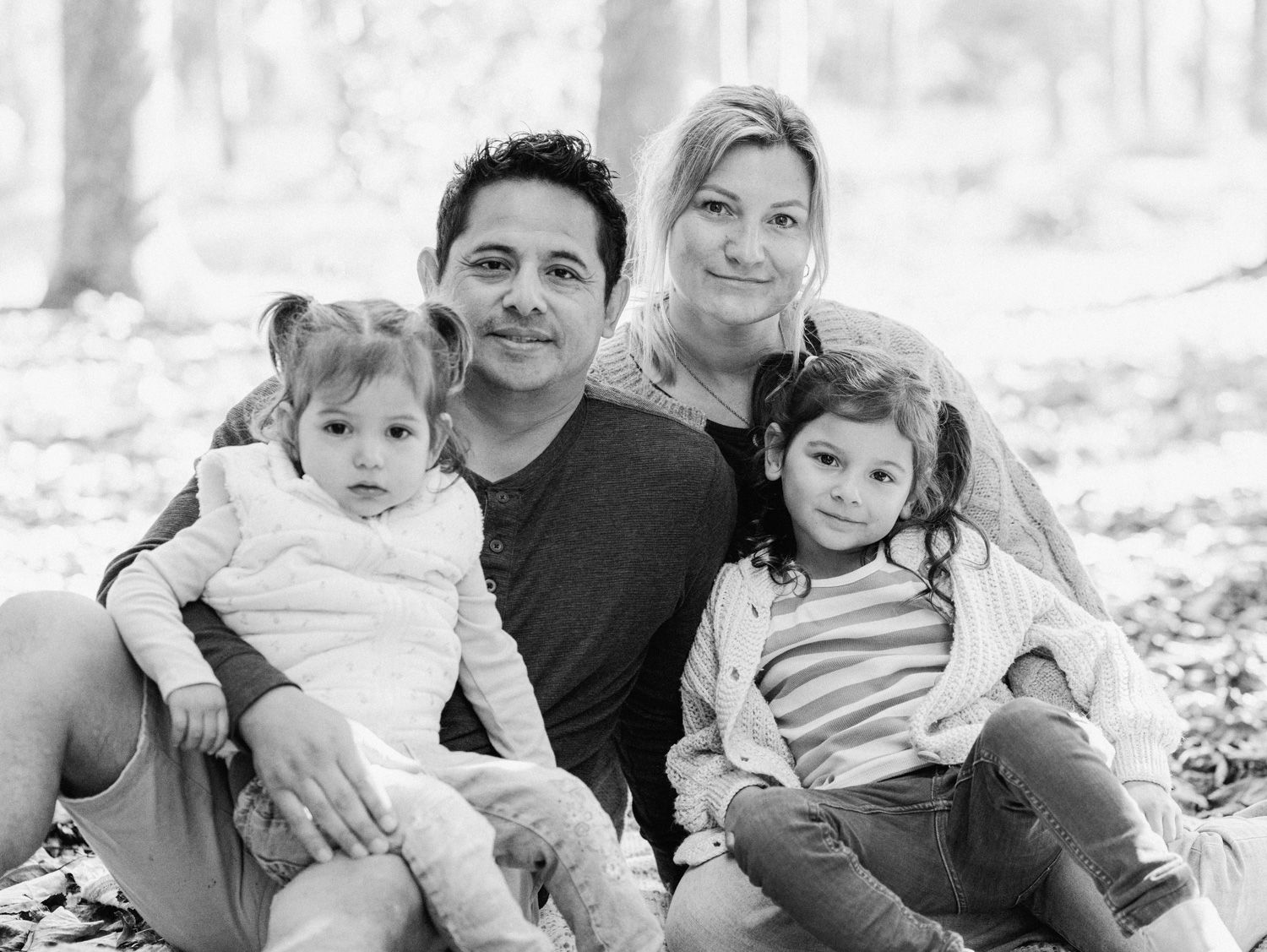 Black and white family portrait of parents with two young daughters sitting outdoors in a wooded setting.