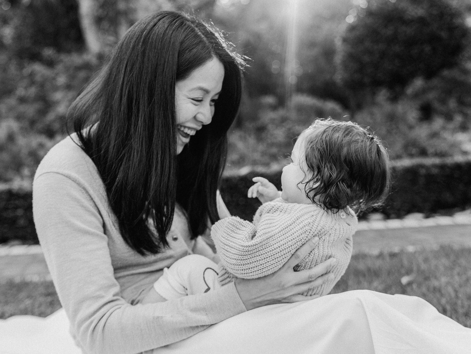 Black and white photo of a smiling Asian mother holding her baby outdoors in a garden setting.