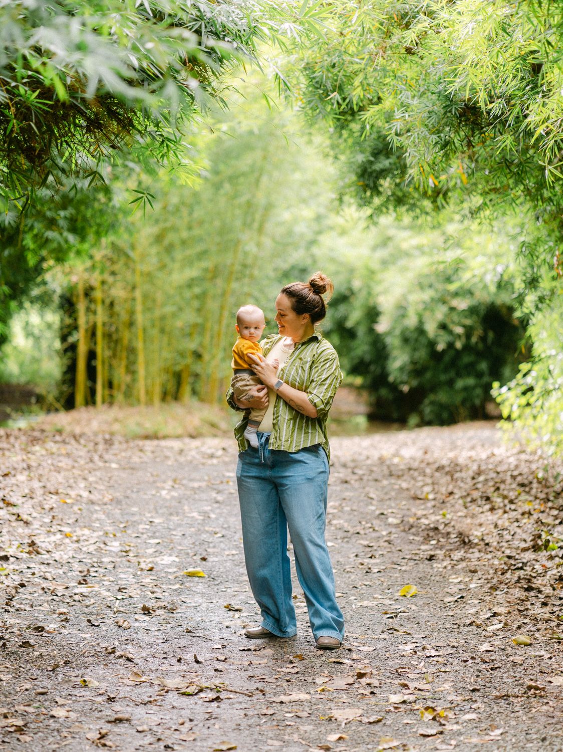 A mother holds her baby on a leaf-covered path surrounded by lush green bamboo trees.