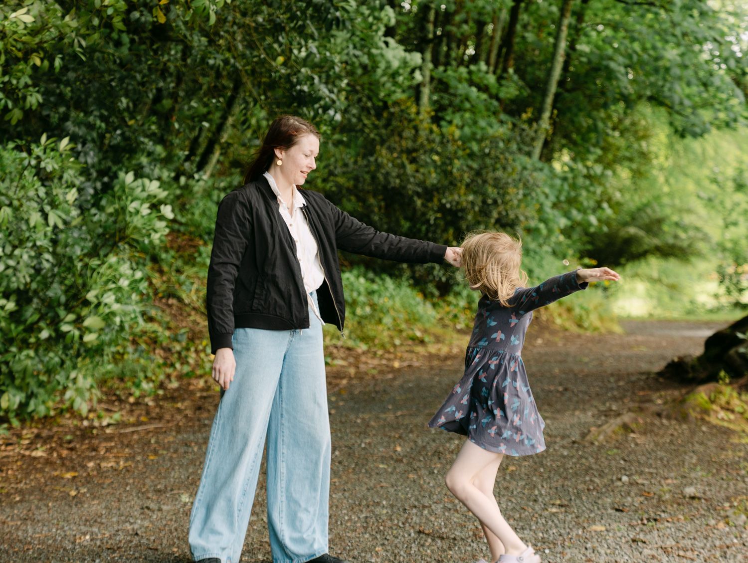 A mother and young girl hold hands while dancing on a path surrounded by lush green trees outdoors.