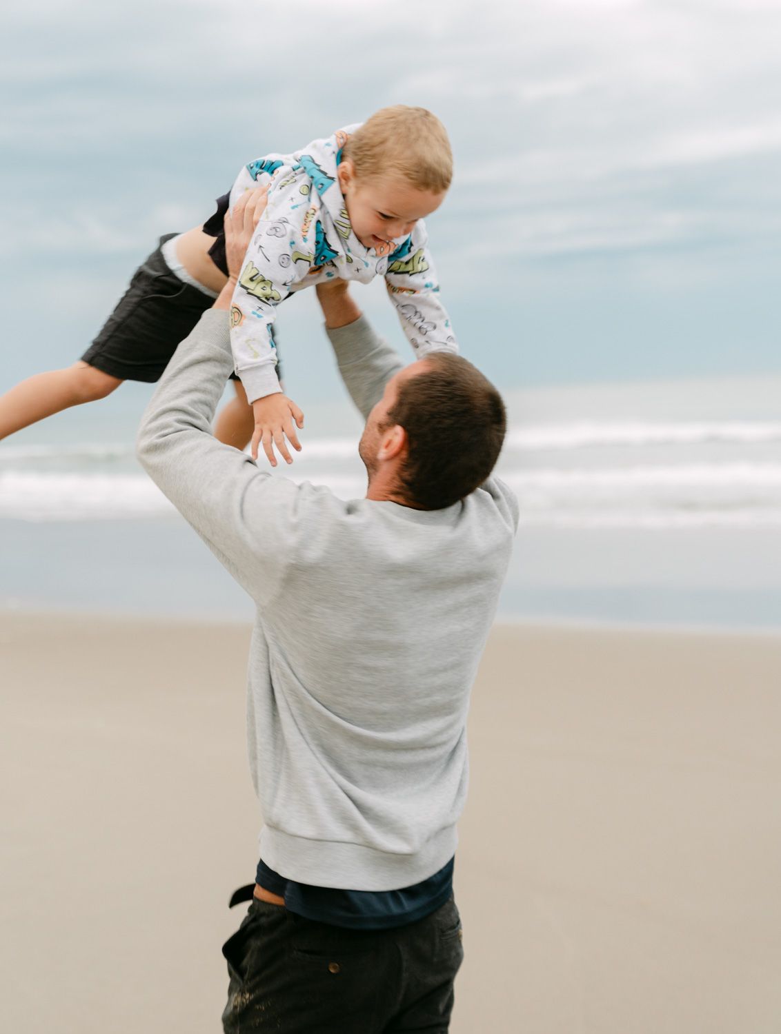 A father joyfully lifts his laughing toddler high in the air on a sandy beach with ocean waves behind them.