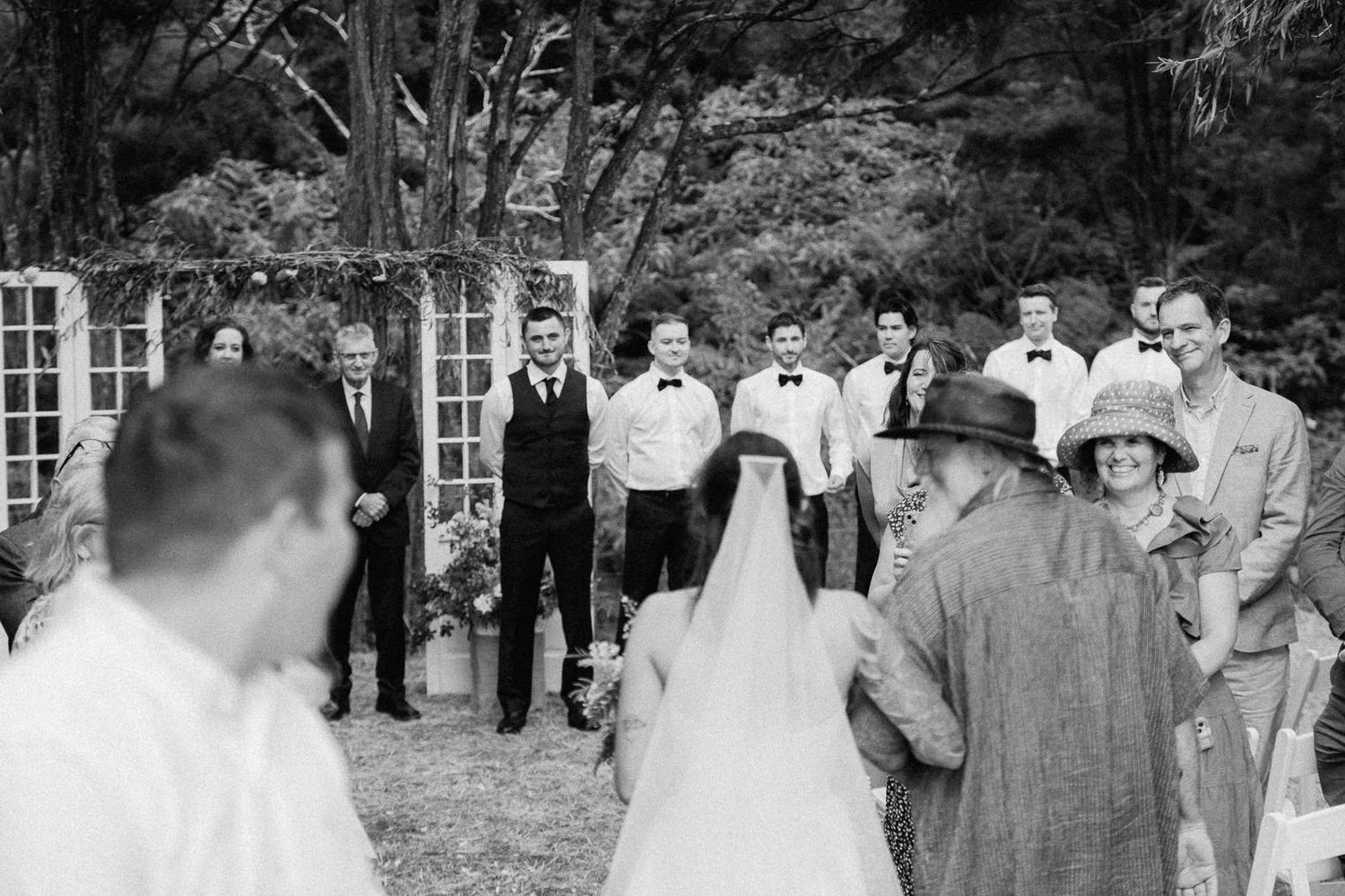 Black and white photo of a bride walking down the aisle toward the groom at an outdoor wedding ceremony.