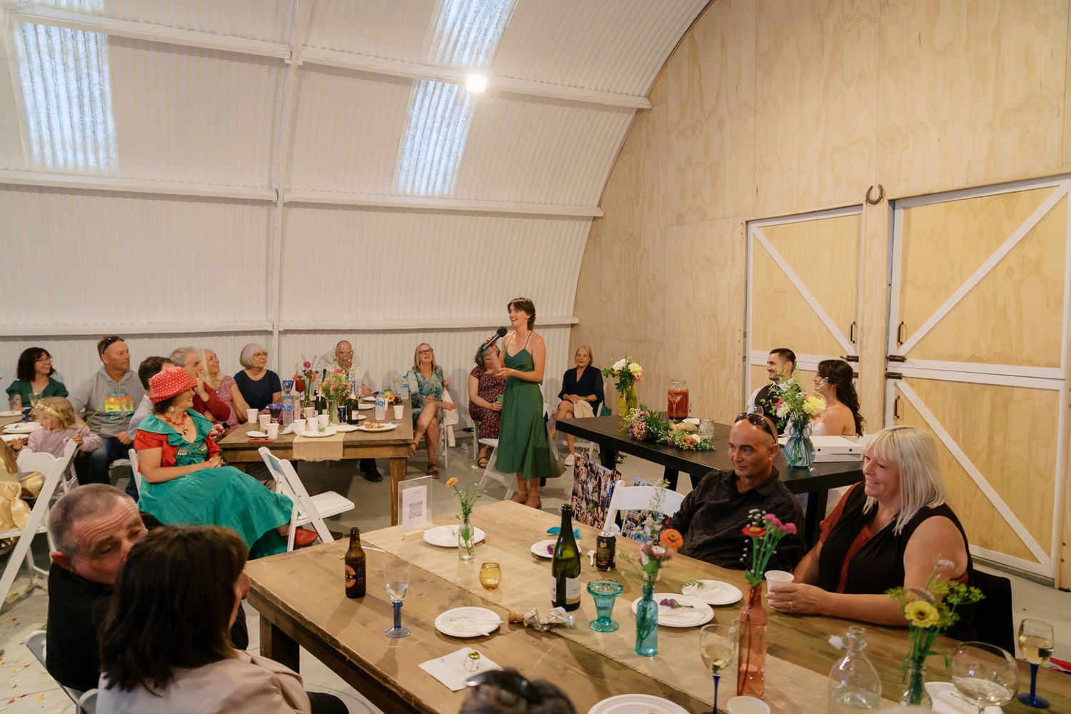 Woman in green dress giving a speech at an indoor wedding reception with guests at long tables.