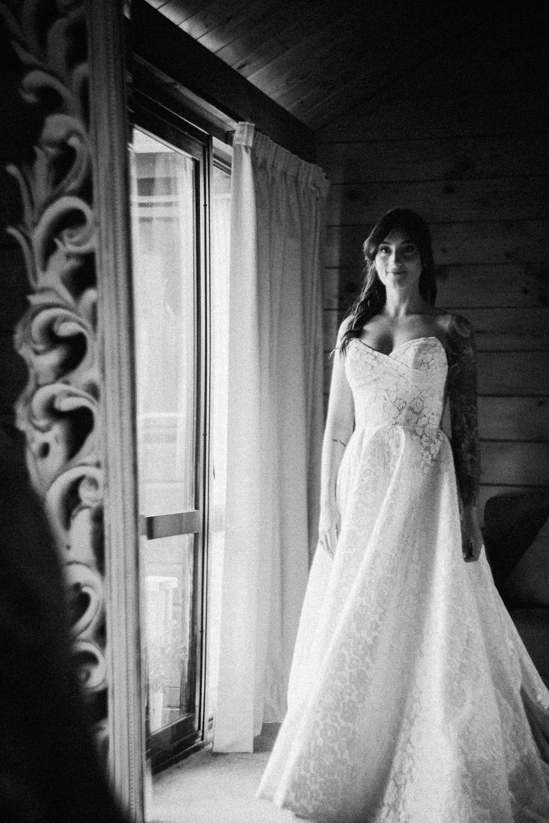 Black and white photo of a bride in a lace strapless wedding gown standing by a window in a wooden room.