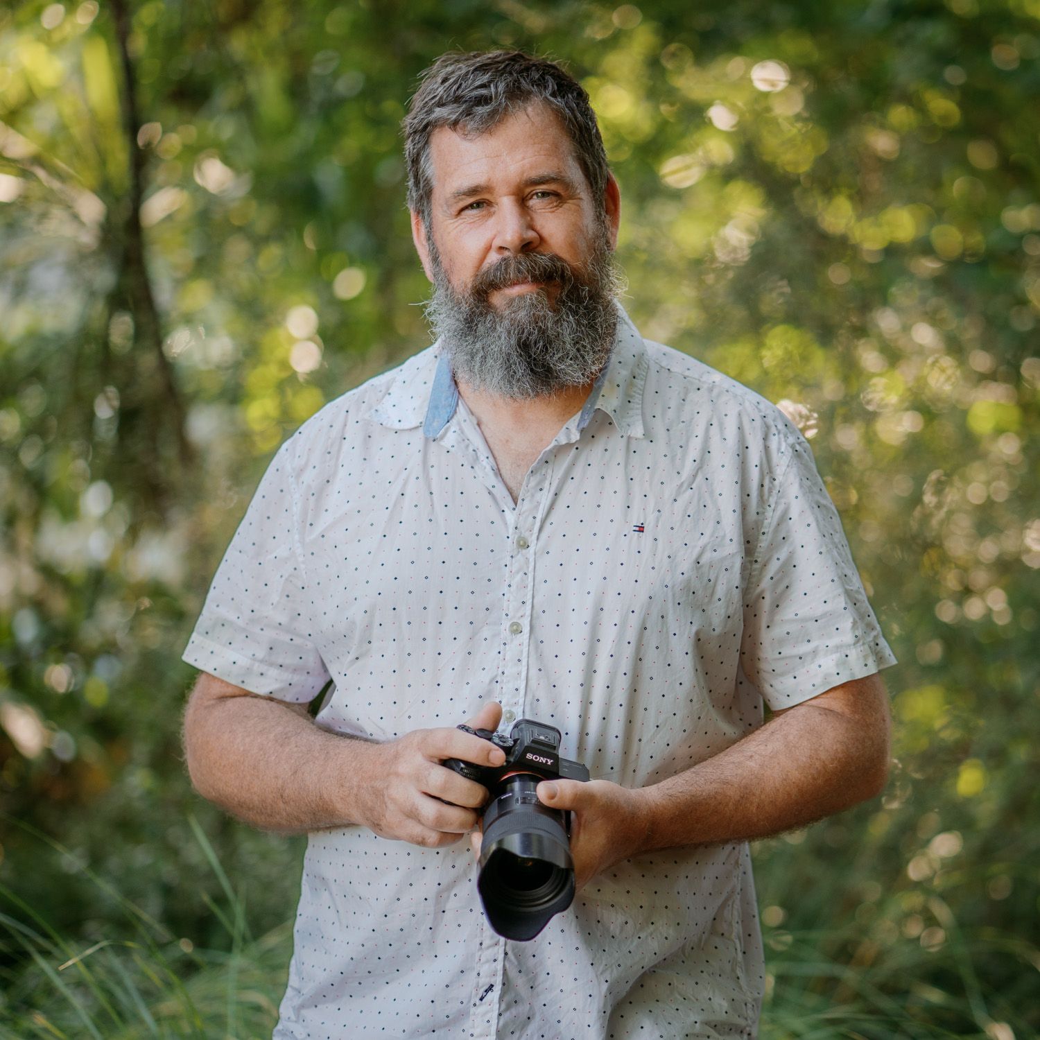 Professional photographer with beard holding camera outdoors in natural forest setting with green foliage background.