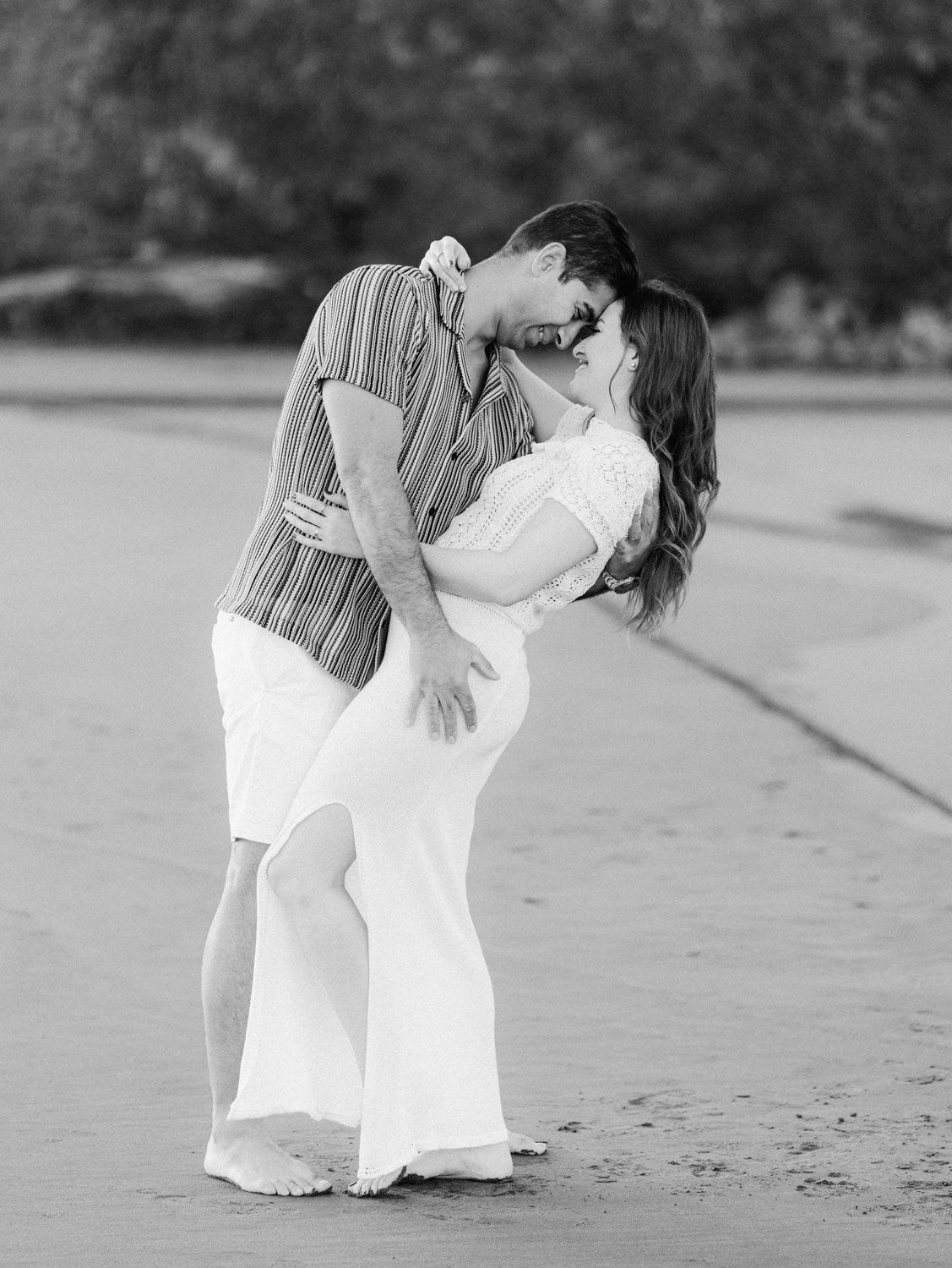 Black and white beach engagement photo of couple sharing romantic dip kiss on sandy shore.