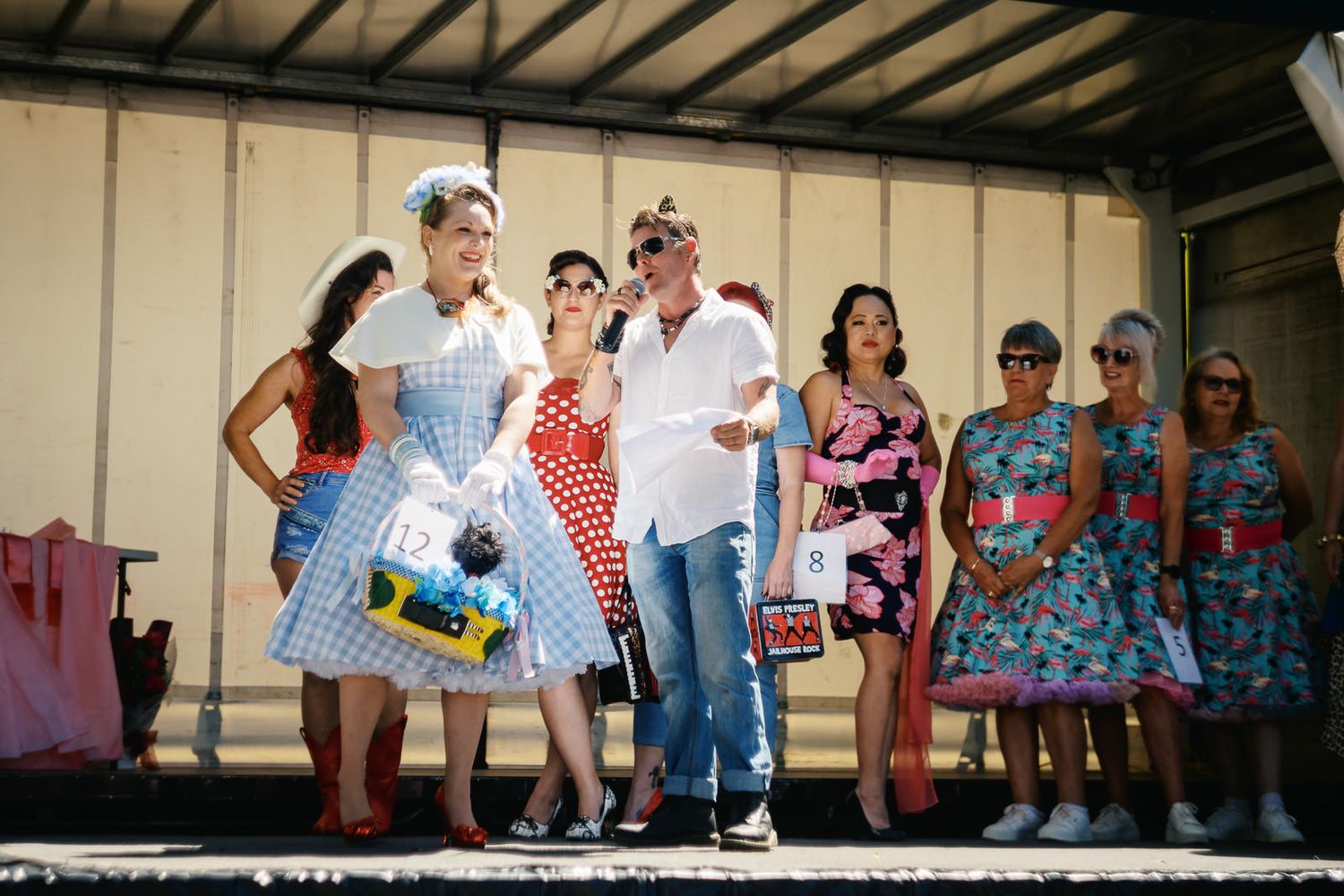 Group of people in colorful 1950s-style outfits posing on a stage at a retro-themed outdoor event.