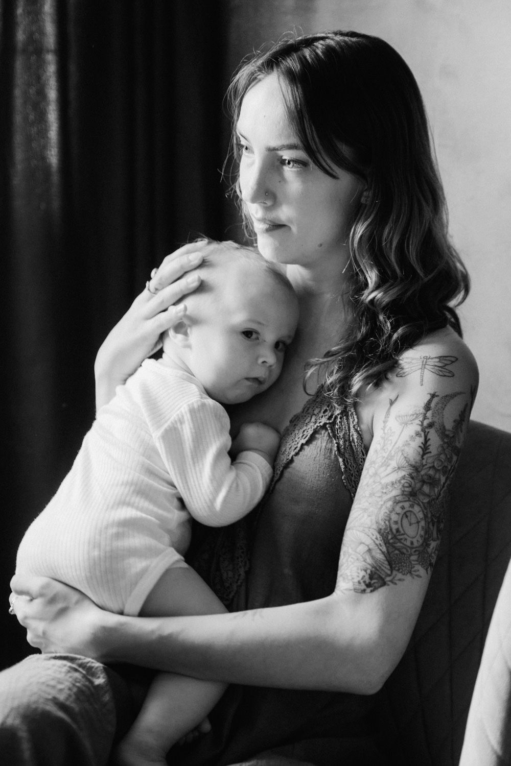 Black and white portrait of a tattooed mother cradling her infant close to her chest while looking away.
