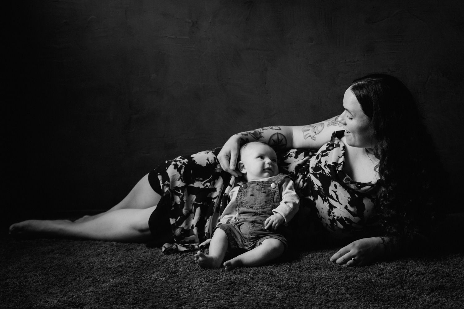 Black and white photo of a mother lying on the floor, tenderly touching her smiling baby in overalls.