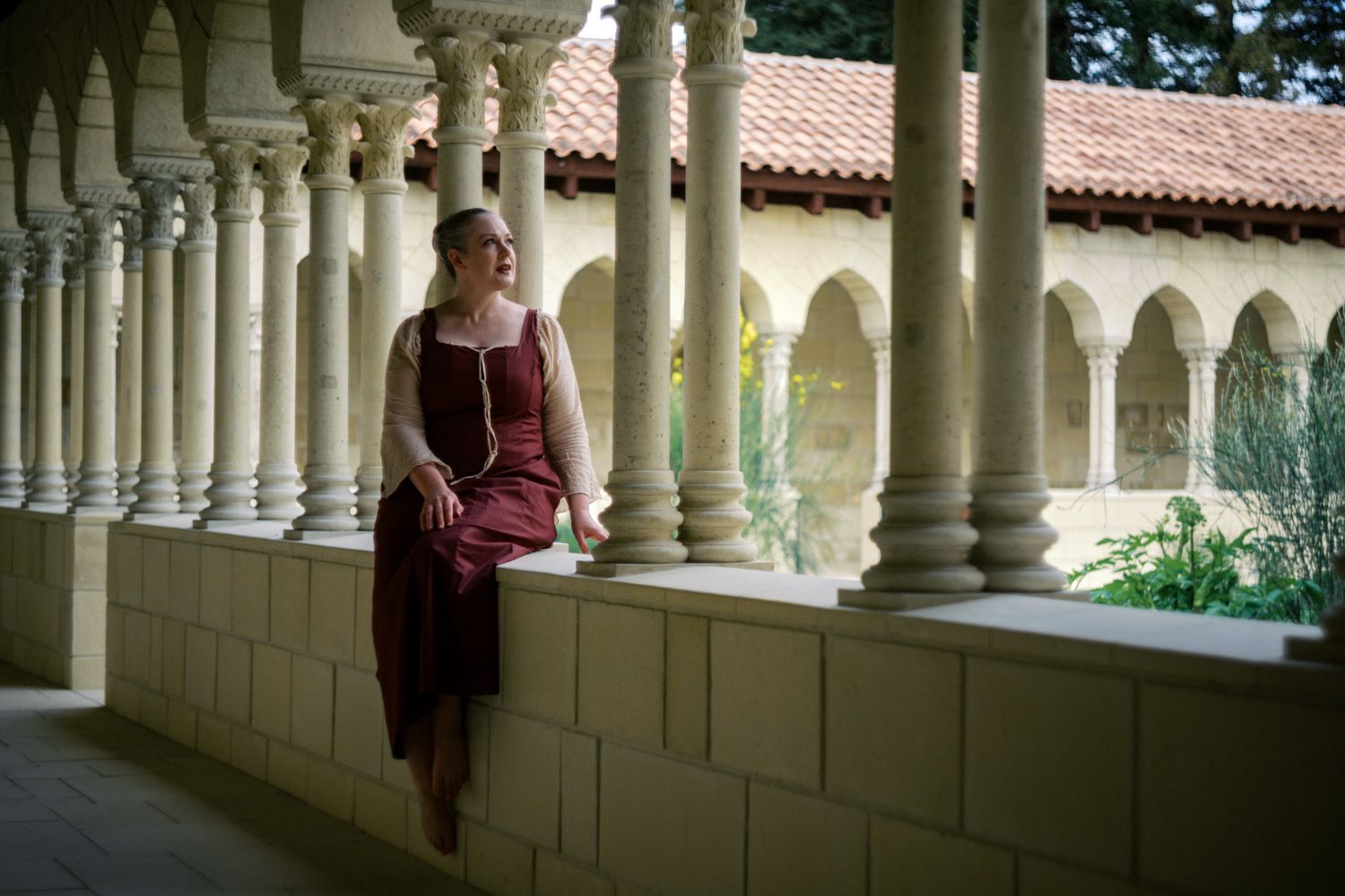 Woman in medieval burgundy dress leaning against stone colonnade in a sunlit Romanesque cloister courtyard.