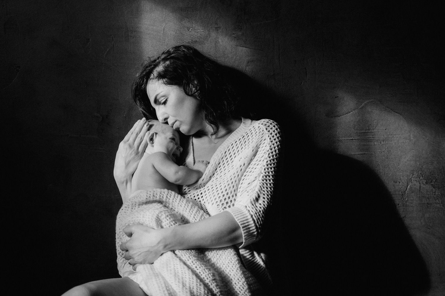 Black and white photo of a mother tenderly holding and kissing her newborn baby in soft dramatic lighting.