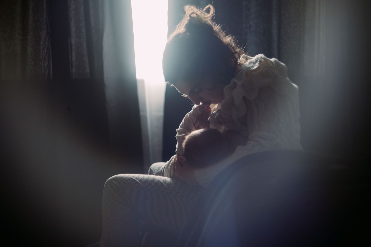 Mother tenderly holding and breastfeeding newborn baby in soft backlit window light.