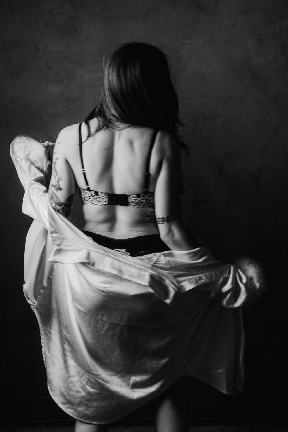 Black and white rear view of tattooed woman in lingerie holding flowing white robe in dramatic studio light.