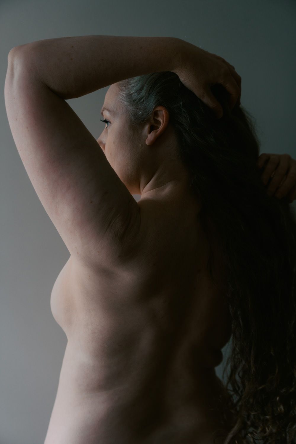 A woman with gray hair poses with arms raised, captured in moody side profile lighting against a neutral background.