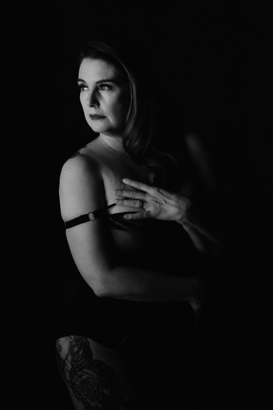 Black and white boudoir portrait of a woman in lingerie with tattoo, posing against dark background.