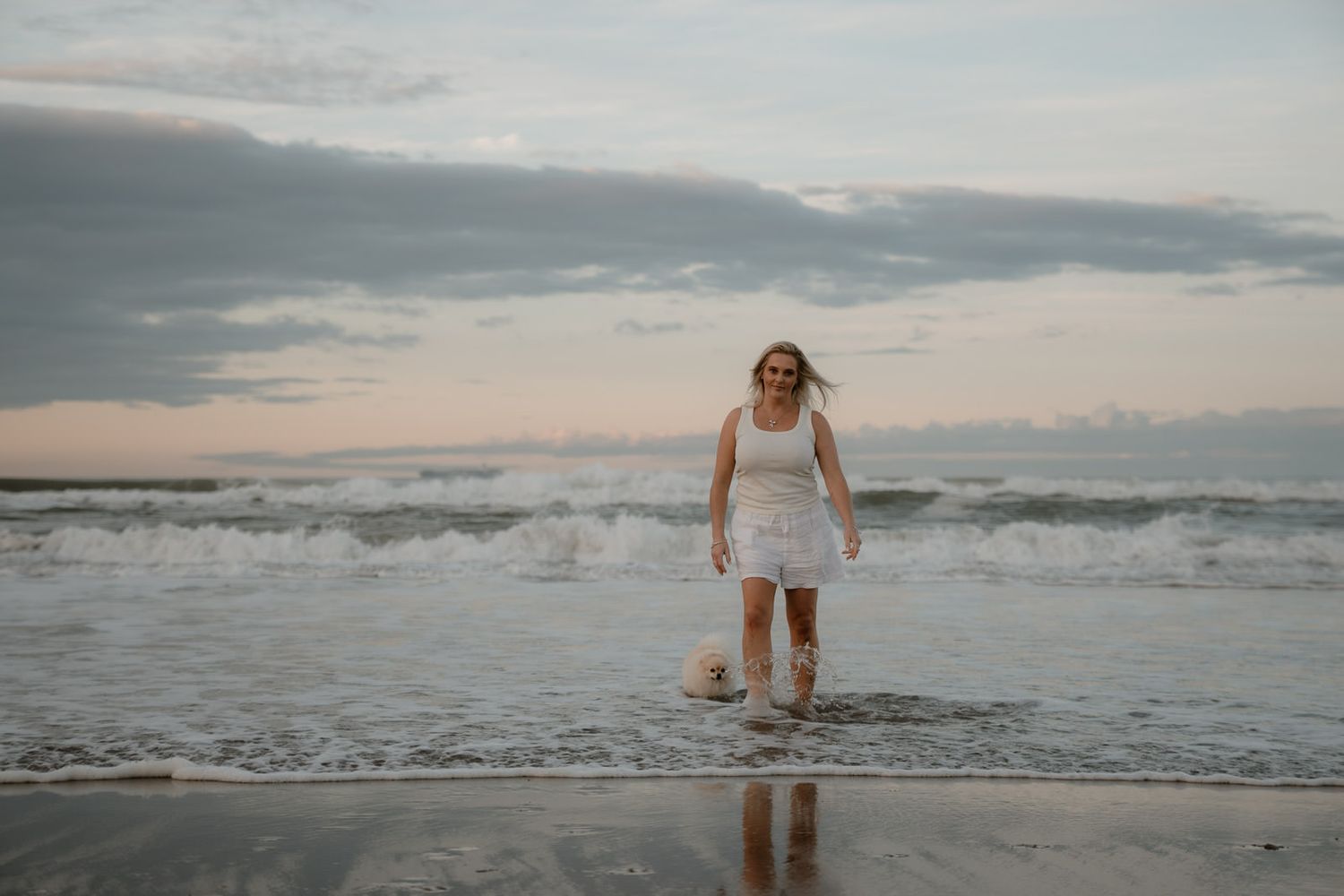 Woman in white dress walking on beach shoreline with ocean waves at sunset under cloudy sky.