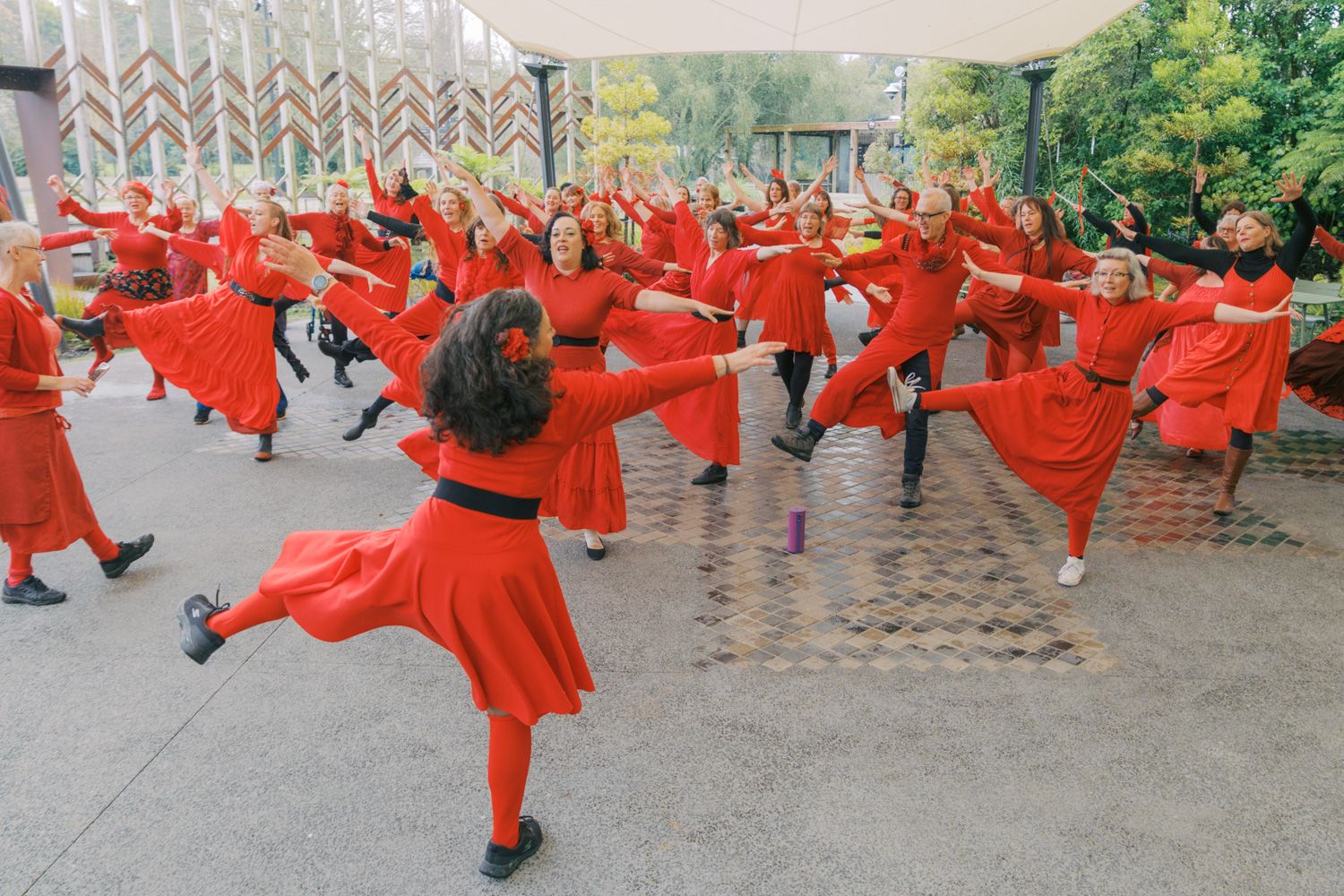 Large group of women in red dresses performing a synchronized outdoor dance routine on a brick plaza.