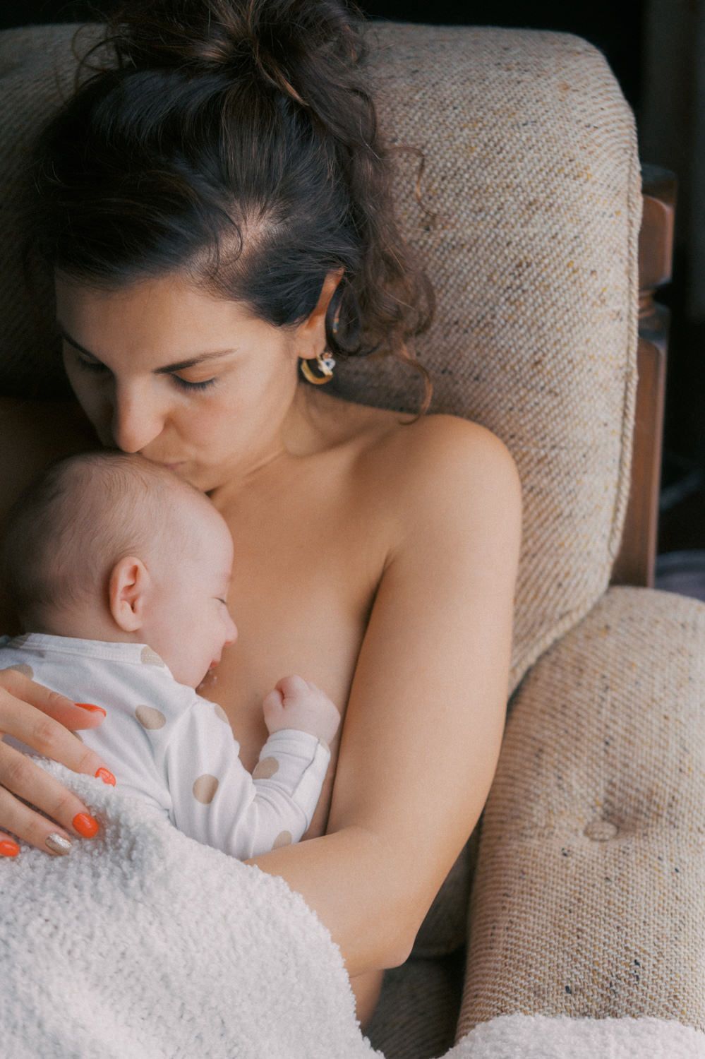A mother tenderly kisses her newborn baby while breastfeeding in a cozy armchair.
