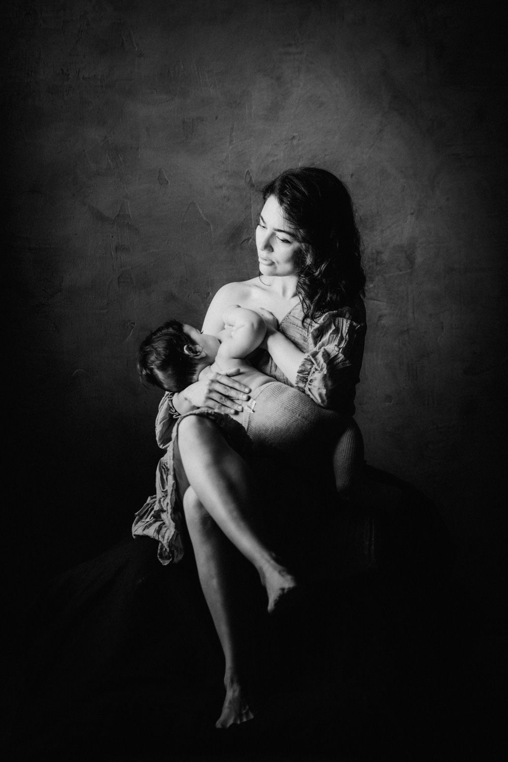 Black and white photo of a mother tenderly breastfeeding her newborn baby while seated against a textured wall.