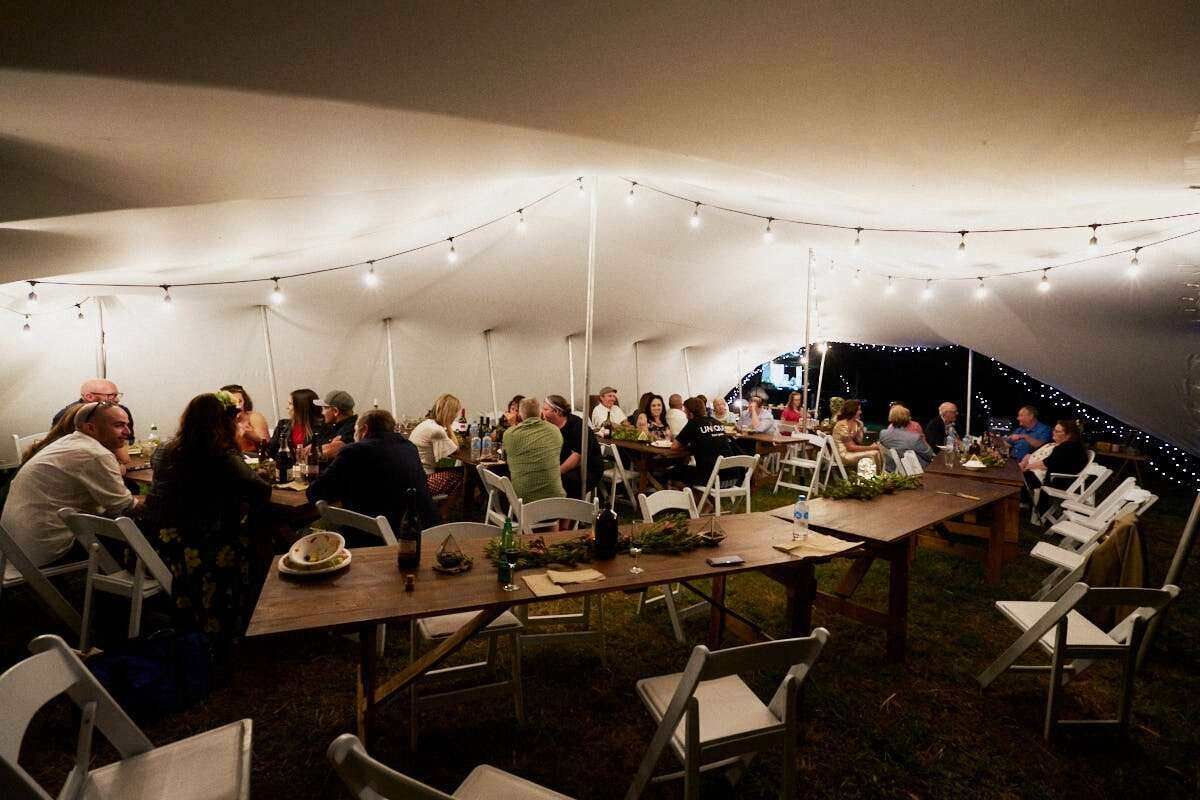 Guests dining under a white tent with string lights at an outdoor evening event with long wooden tables.