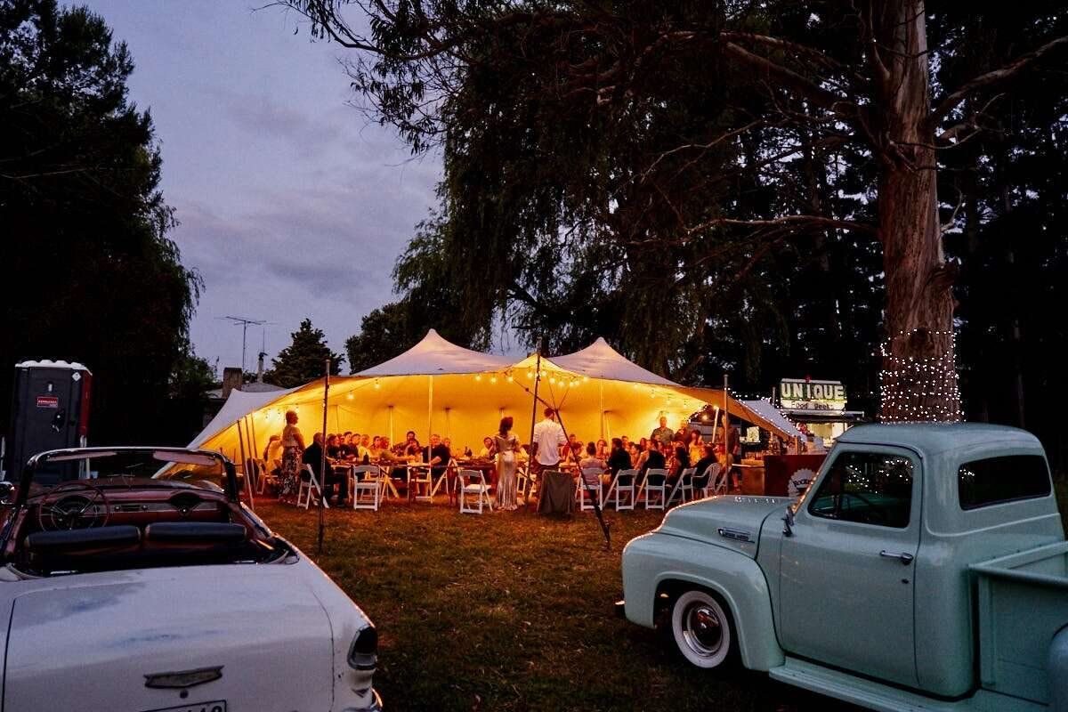 Glowing sailcloth tent at dusk surrounded by vintage classic cars and string lights at an outdoor evening event.