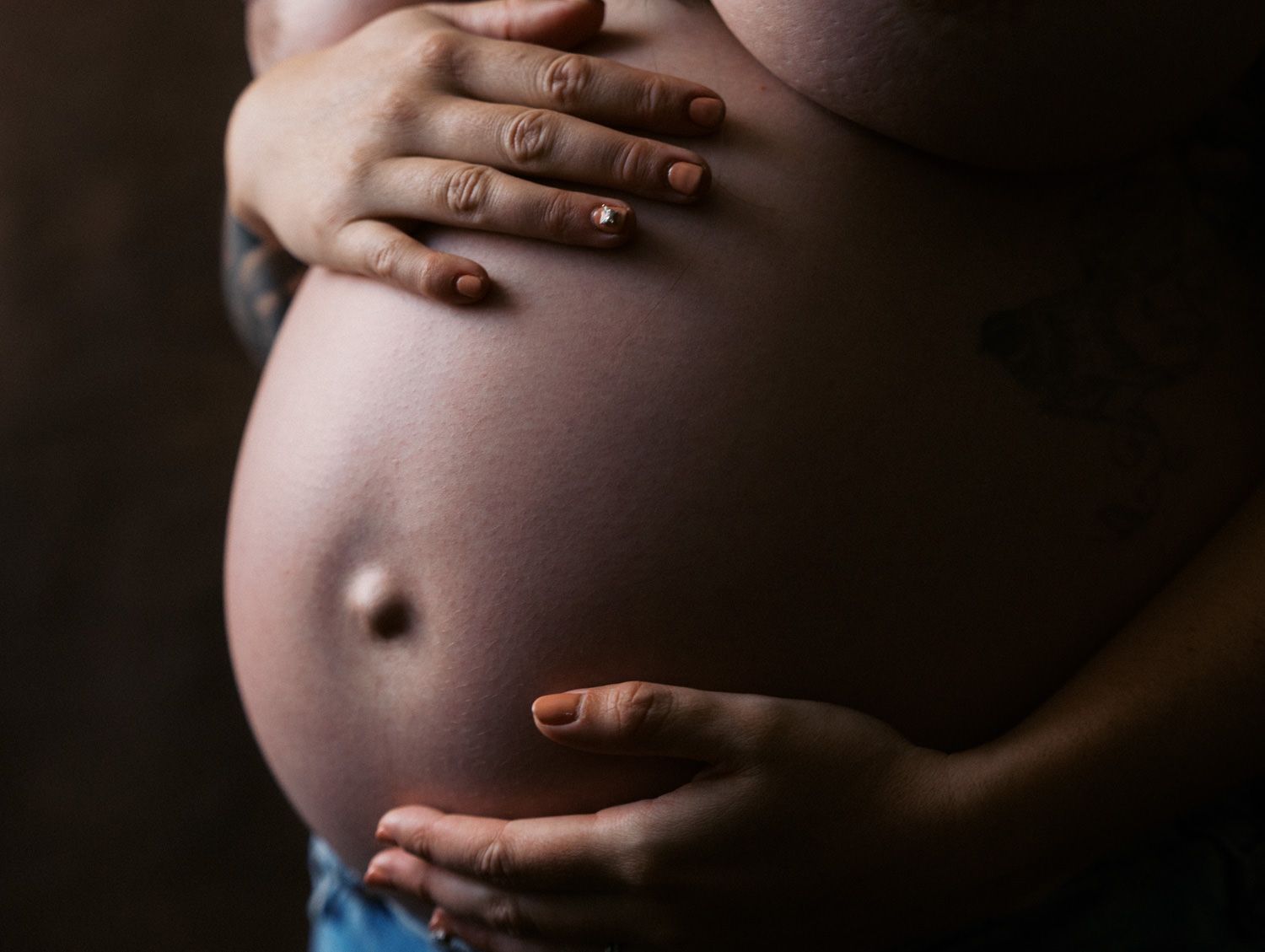 Close-up of a pregnant woman cradling her bare baby bump with both hands, showing a tattoo on her side.