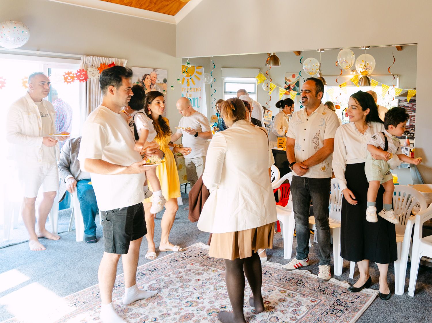 Families gather indoors at a cheerful celebration with balloons and decorations in a bright, festive room.