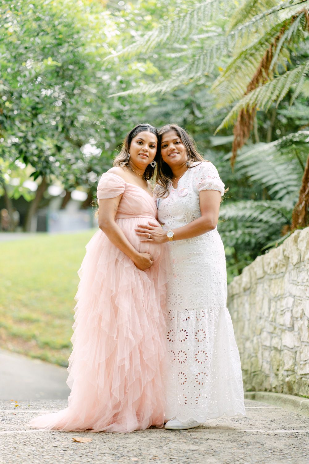 Two women in elegant pink tulle and white lace gowns pose together outdoors in a lush green garden setting.