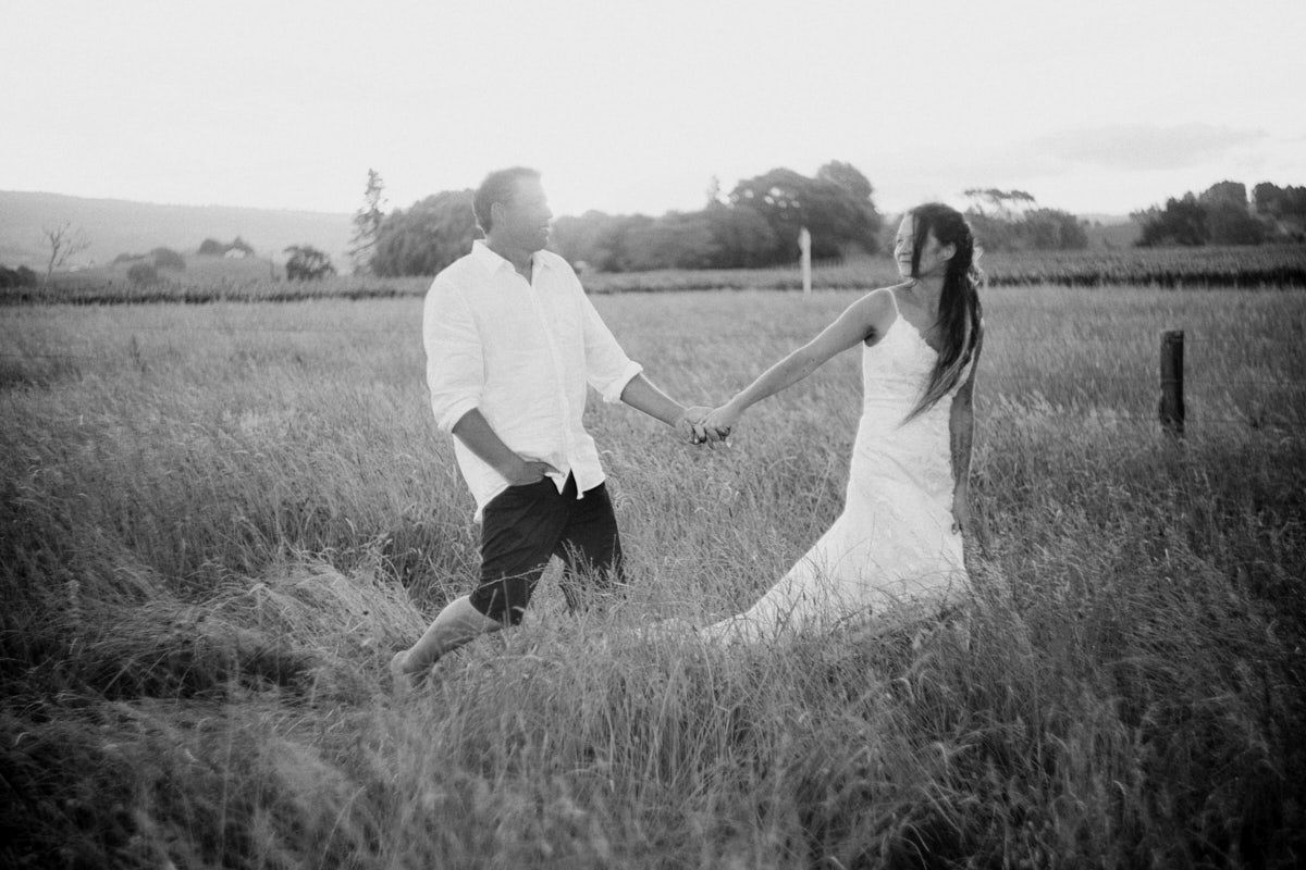 Black and white photo of a couple holding hands walking through a lush field at sunset.