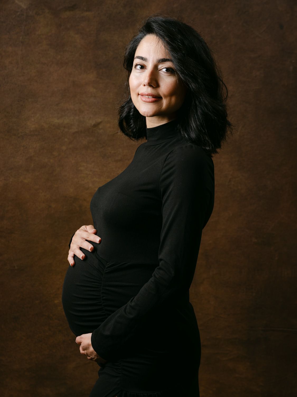 Pregnant woman in black turtleneck dress cradling baby bump, posing sideways against brown studio backdrop.
