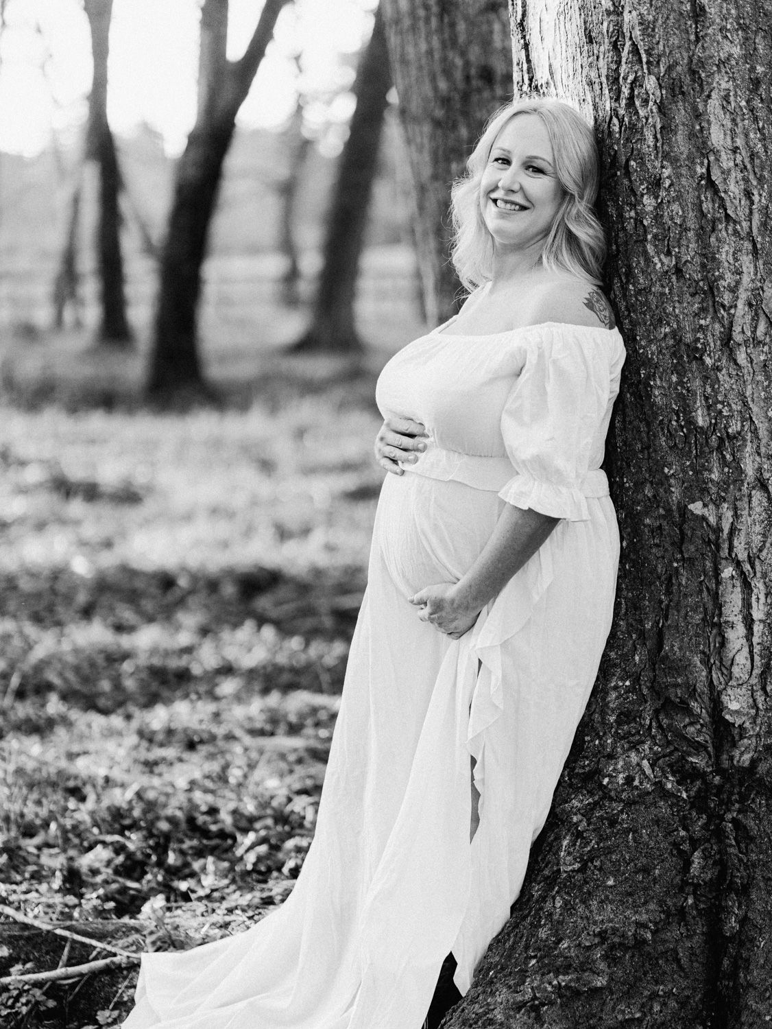 Pregnant woman in white off-shoulder gown leaning against tree in black and white outdoor maternity portrait.