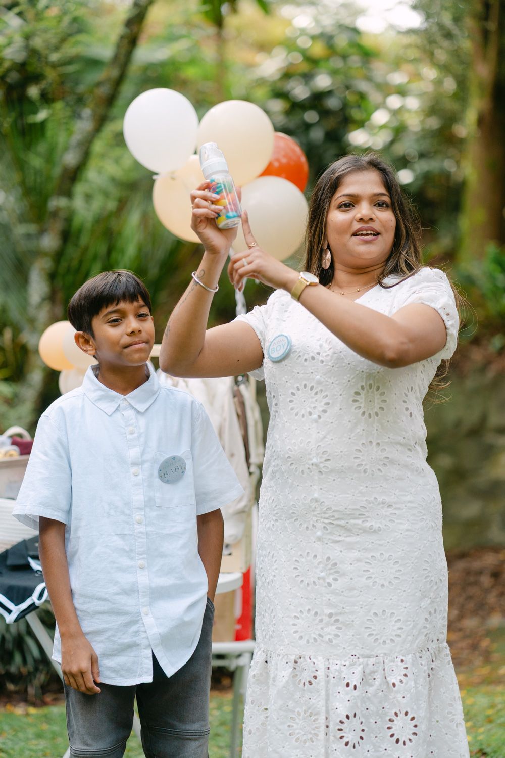 Woman in white dress and boy at outdoor party with balloons, holding a small decorated bottle.