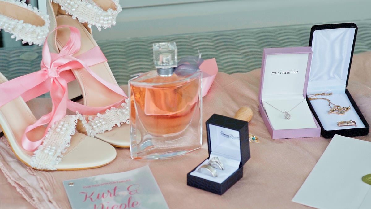 Bridal flat lay with pearl heels, Lancome perfume, diamond ring, pink jewelry box, and gold necklace on a pink surface.