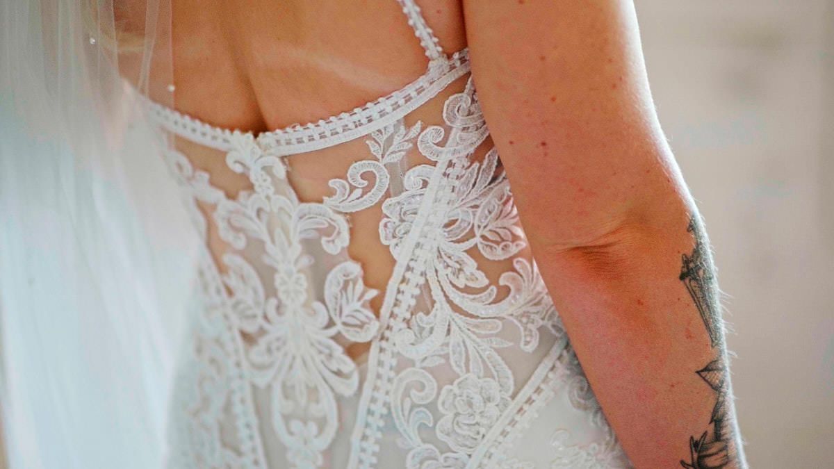 Close-up of intricate lace wedding dress back with pearl details and bride's arm tattoo visible.