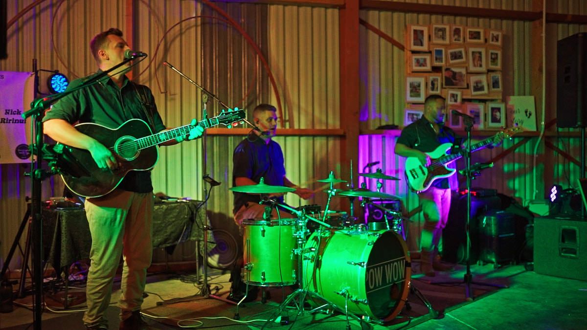 A live band performs on a green-lit stage with a guitarist, drummer, and bassist in a rustic venue.