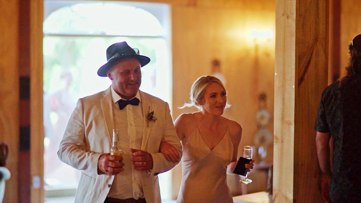 Bride and groom entering wedding reception holding drinks, groom in white suit and hat, bride in elegant white gown.