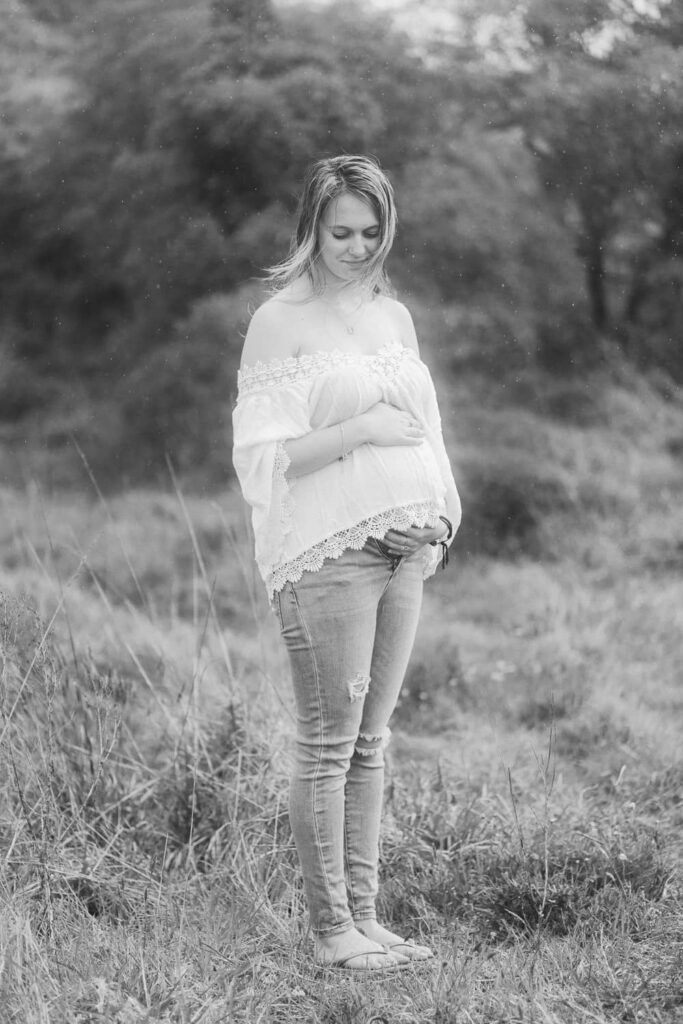 Black and white maternity photo of a pregnant woman in white off-shoulder top and jeans, cradling her baby bump outdoors.