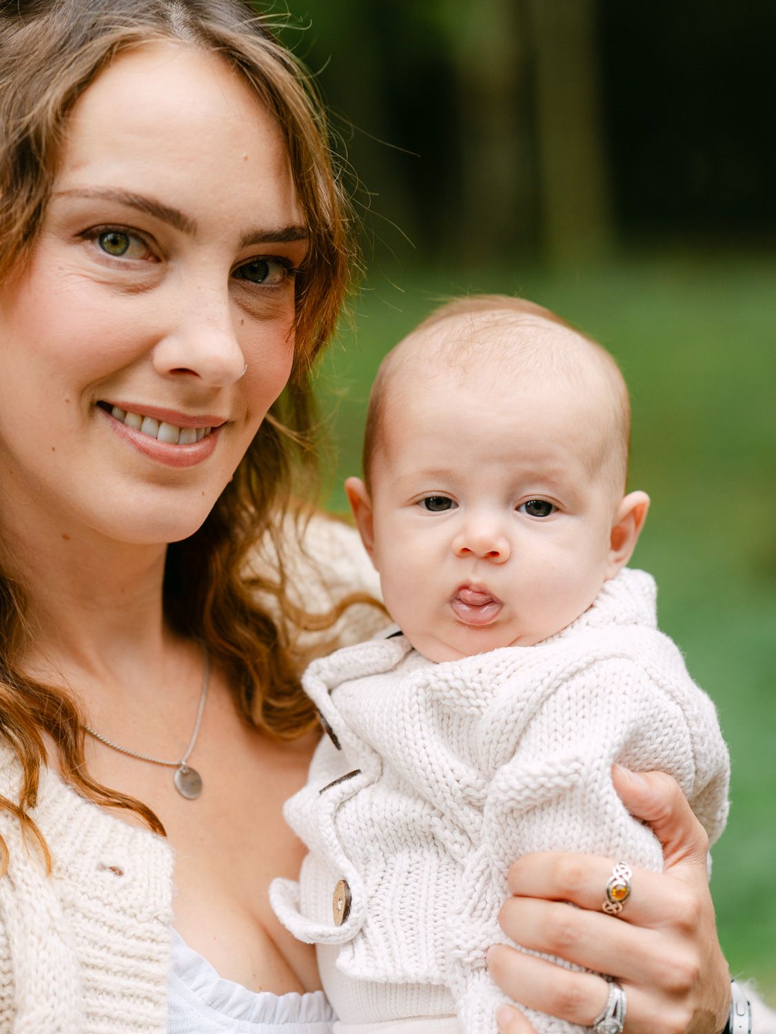 A smiling young woman with brown hair holds a cute baby in a white knit outfit outdoors in a green setting.