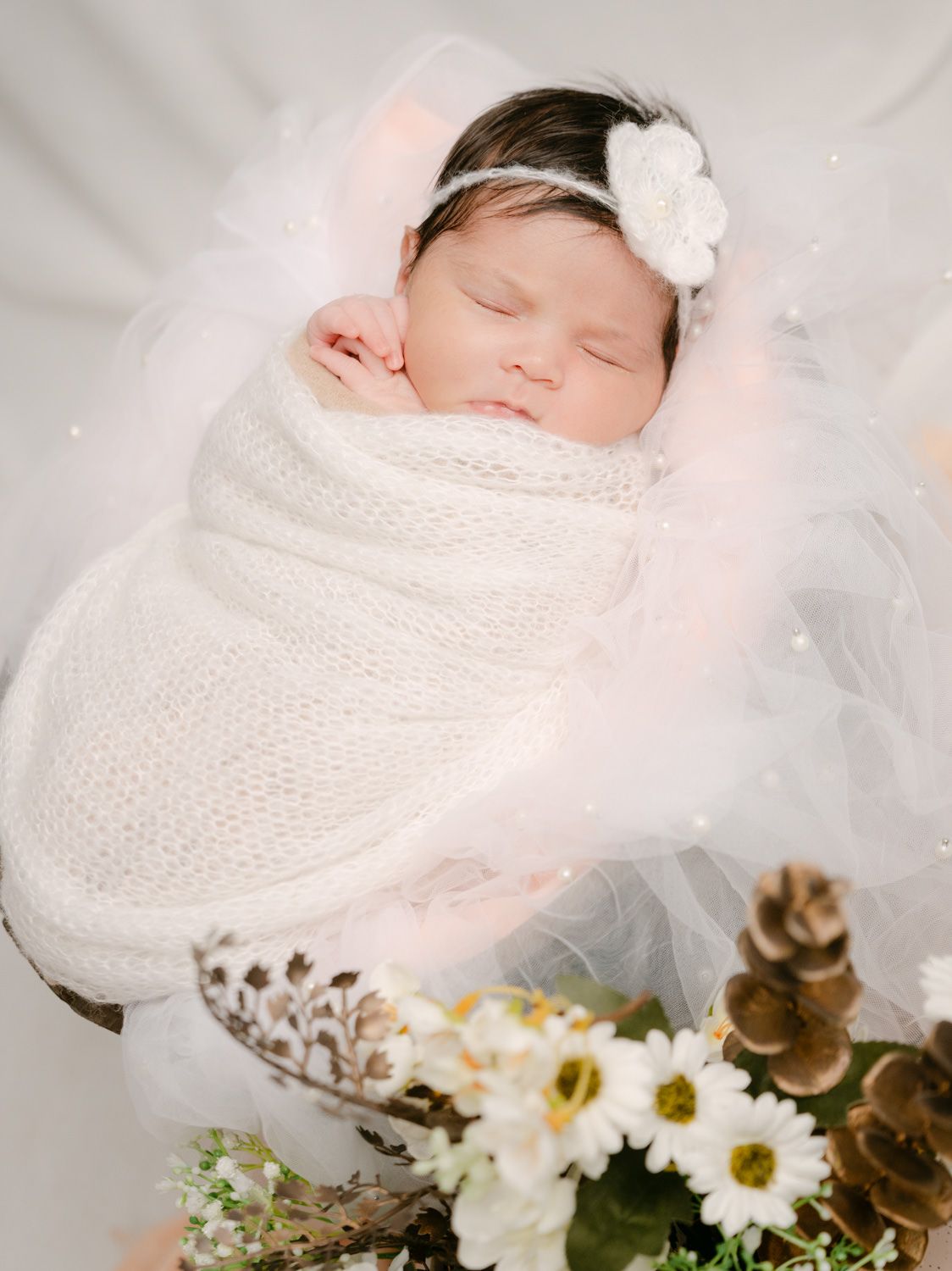 Sleeping newborn baby swaddled in white knit wrap with floral headband, surrounded by daisies and pinecones.