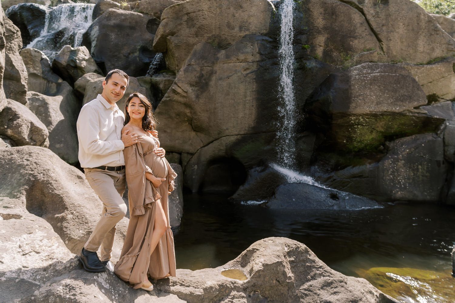 Couple posing on rocks in front of a waterfall, woman in flowing beige dress, man in white shirt, outdoor maternity photo.